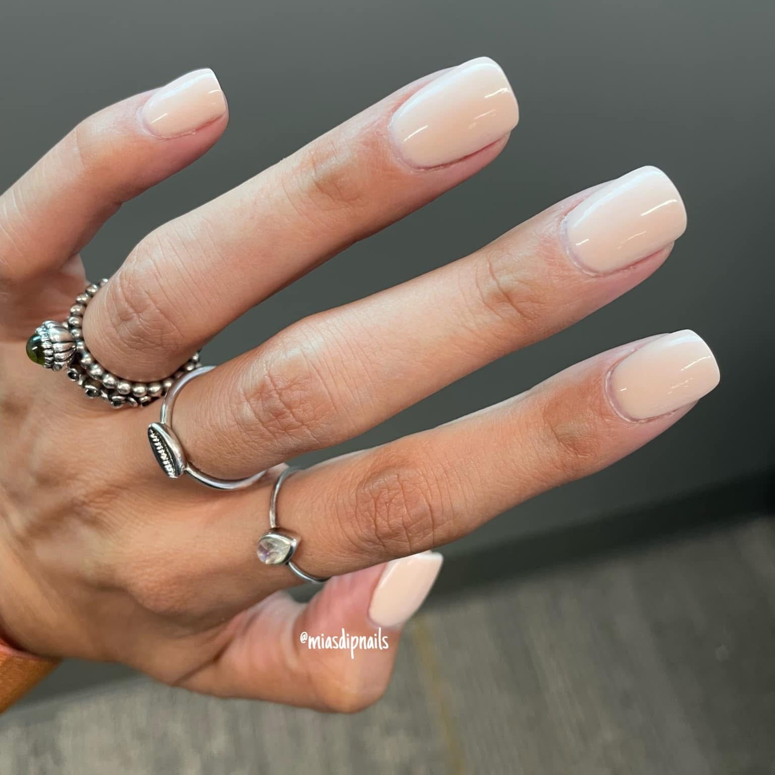 Manicured hand with glossy beige nude nails and silver rings on fingers.