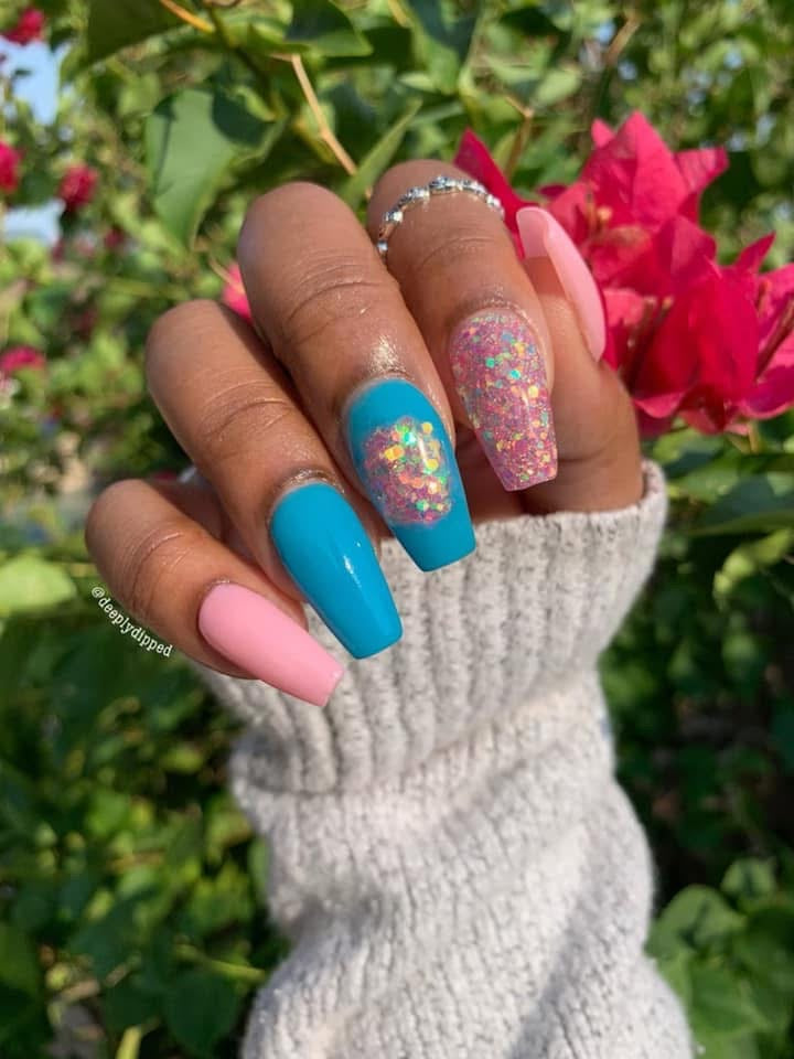Manicure with pink, bright blue, and multicolor chunky glitter nails on hand wearing light gray knit sleeve, outdoors with.