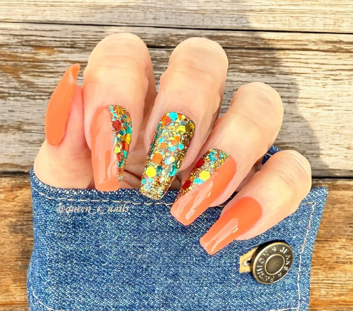 Long almond-shaped nails painted in solid peach orange with two accent nails decorated with colorful hexagonal glitter on a.