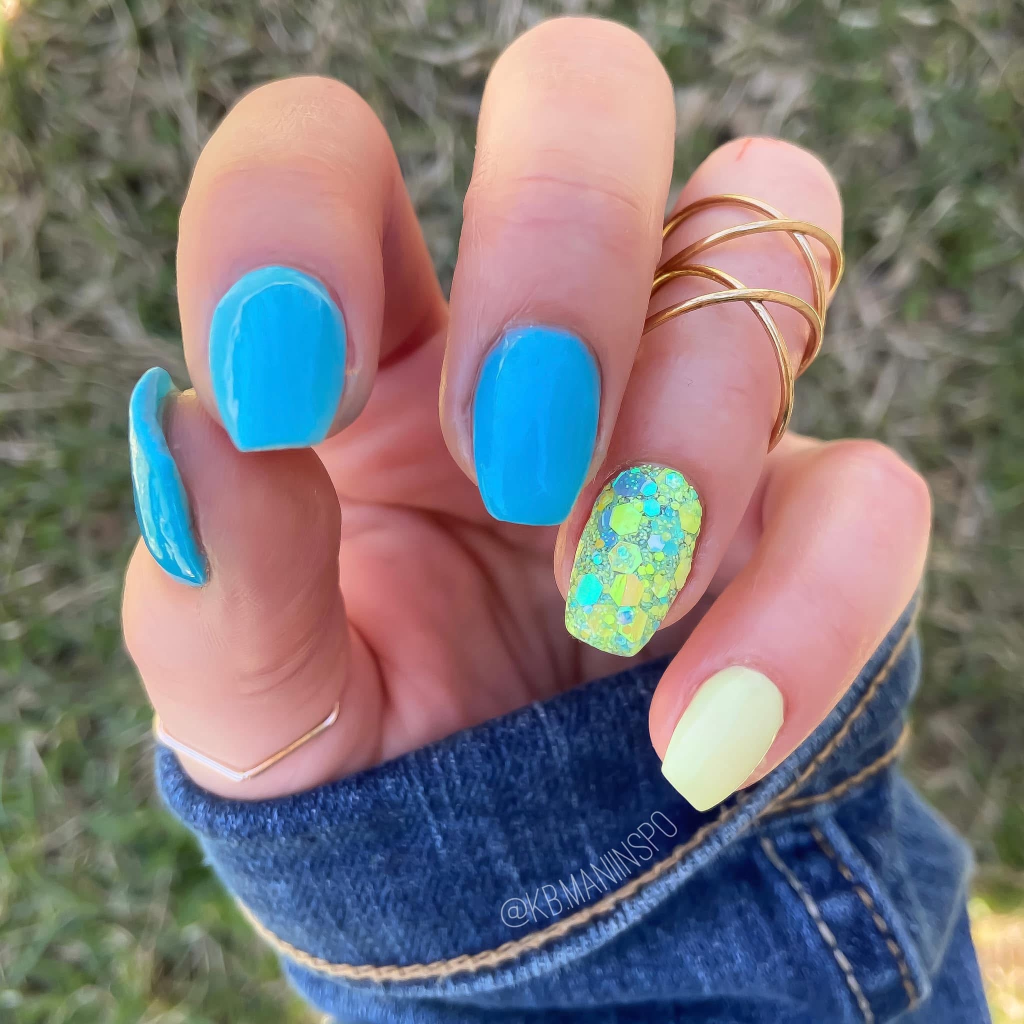 Hand with blue dip powder nails, one accent nail with green glitter, and one pale yellow nail, wearing a gold ring and denim .