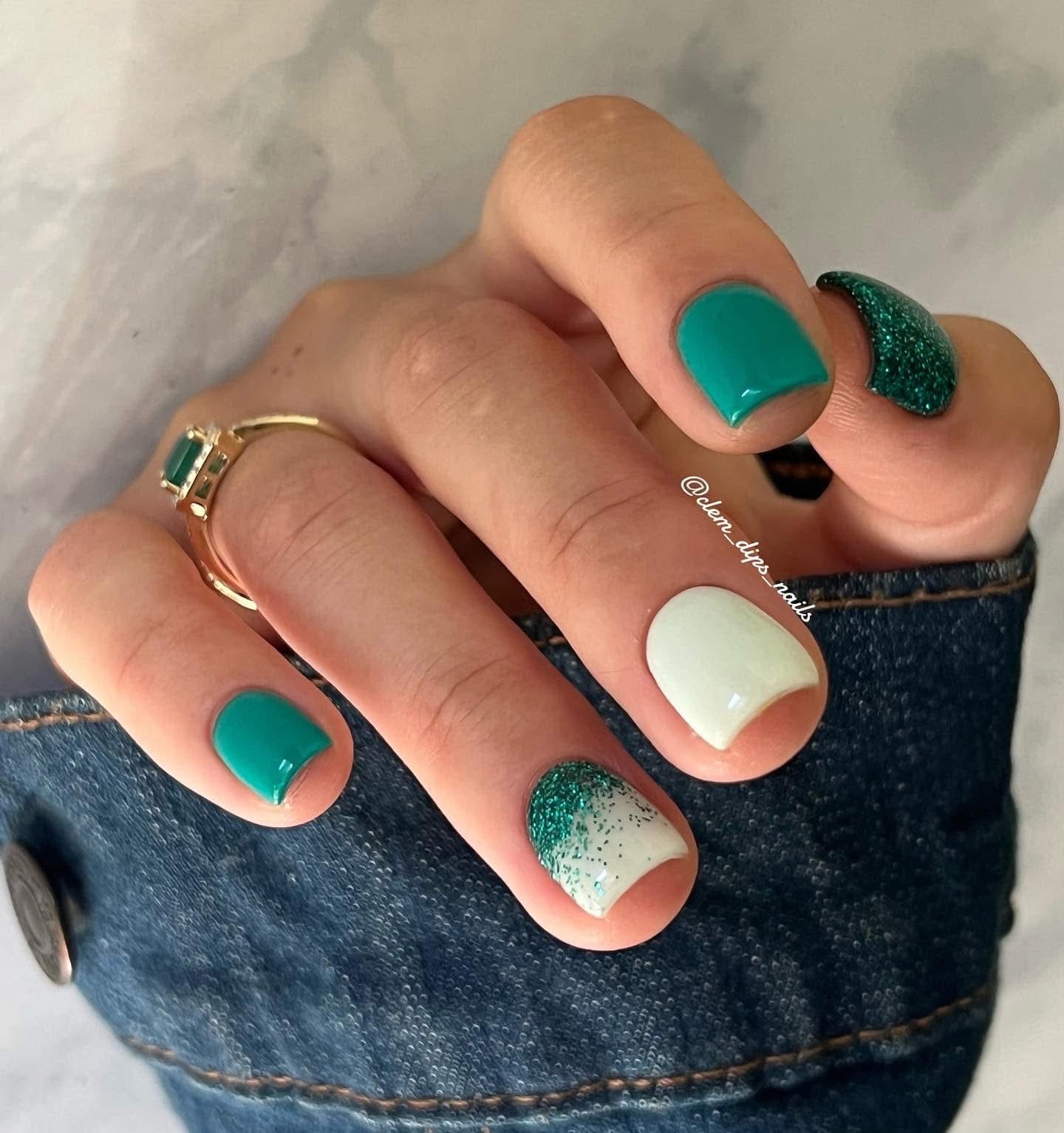 Manicure with teal green solid polish on four nails, white accent nail with teal glitter gradient, hand resting on denim.