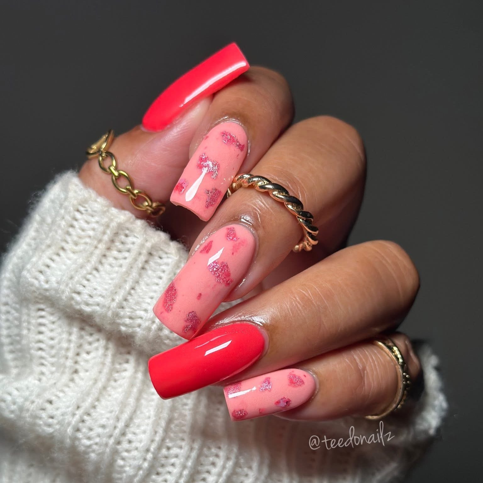 Hand with pink and red dip powder nails and gold rings on a dark background