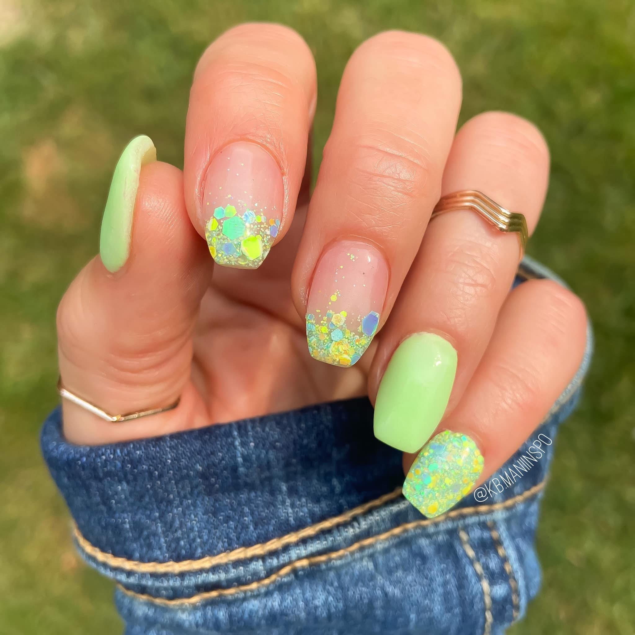 Natural nails with pastel green polish and chunky multicolor glitter floral nail art on ring and pinky fingers.