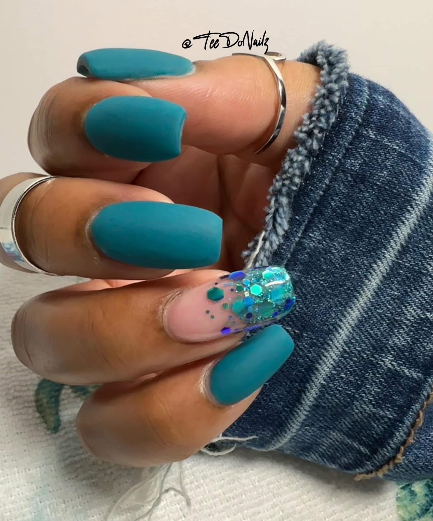 Matte teal dip powder nails with one glitter accent nail decorated with blue and silver flakes.