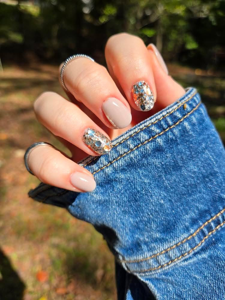 Hand with beige nude solid dip powder nails, two nails decorated with silver foil accents, wearing silver rings, holding.