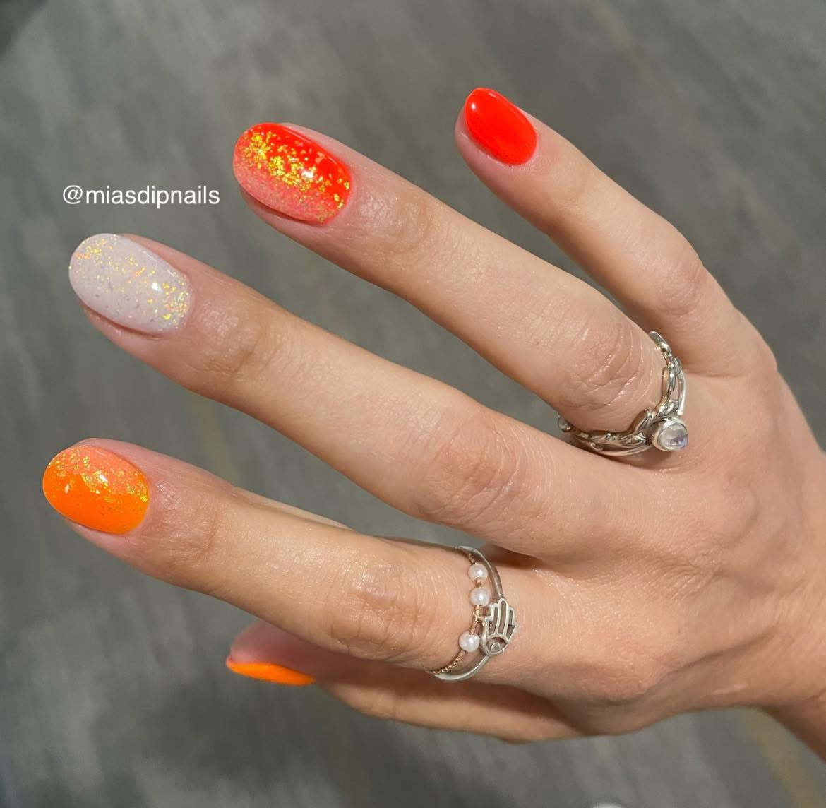Bright neon orange and glowing orange dip powder nails with iridescent glitter accent on a natural nail base.