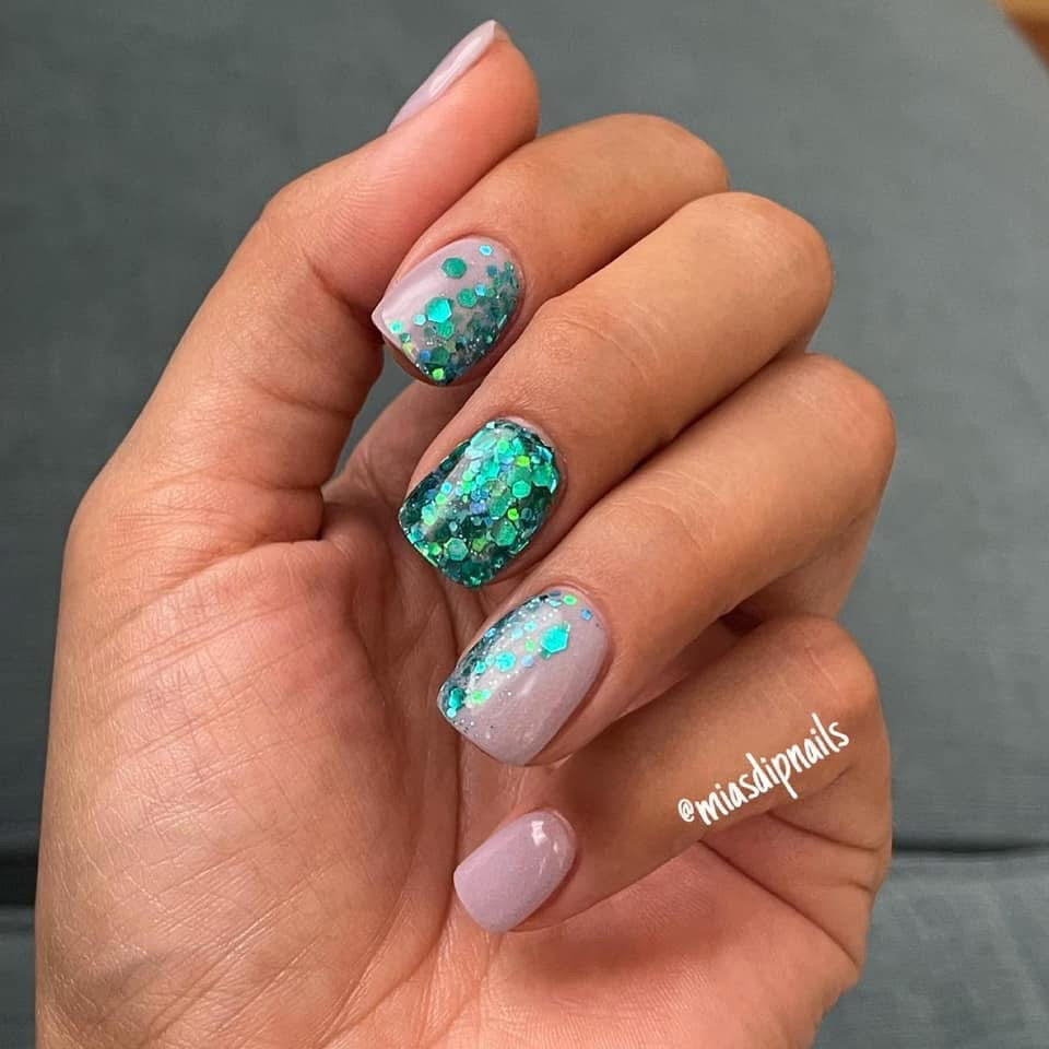 Hand with lilac dip powder nails accented by turquoise and green glitter on index and ring fingers.