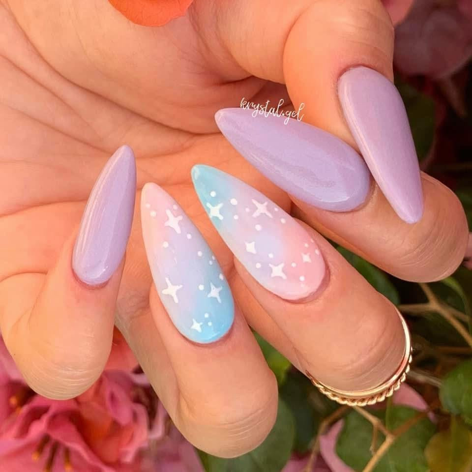 Long almond-shaped nails with lilac polish and two nails featuring pastel pink and blue gradient with white star and cross.