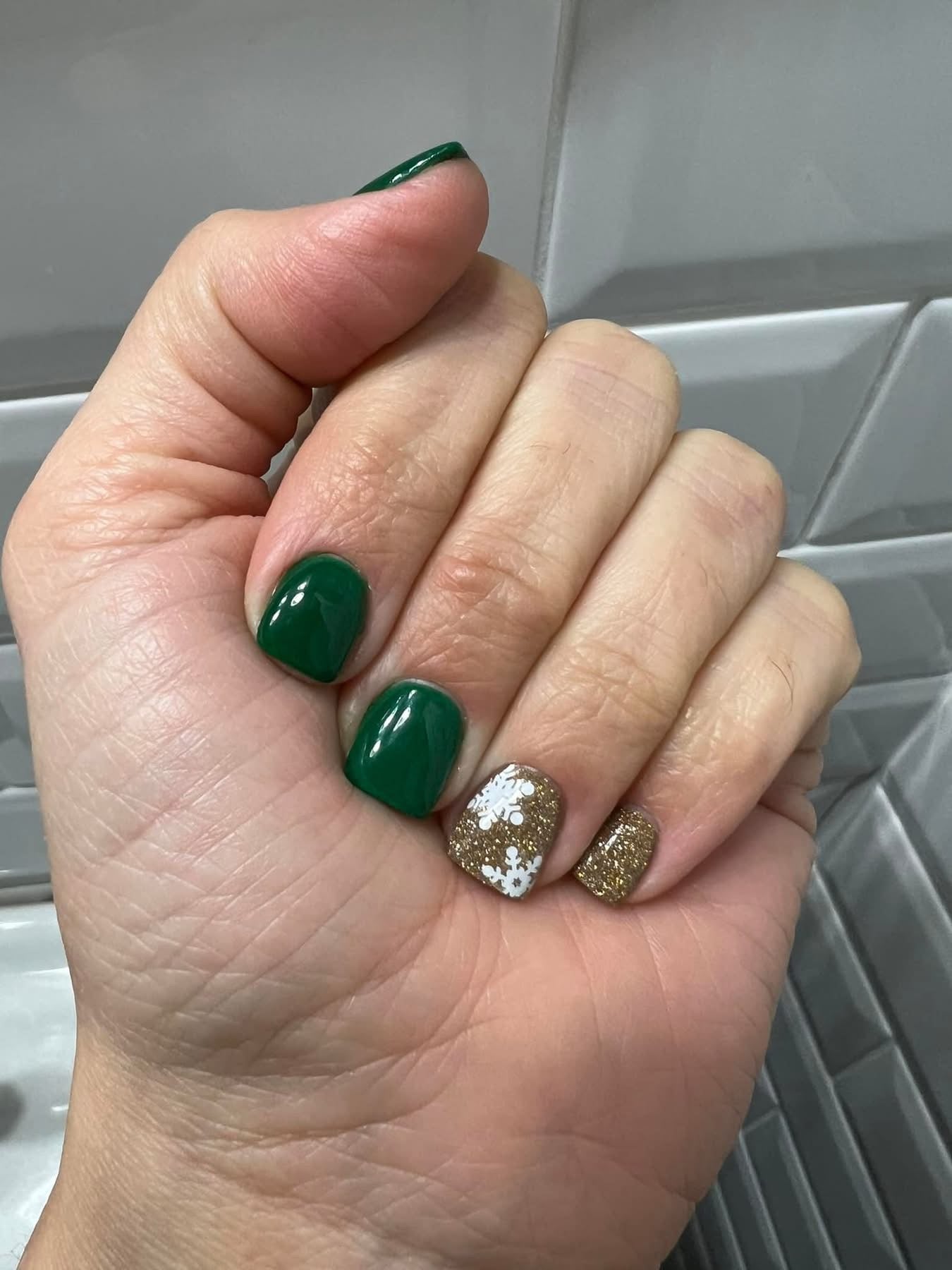 Dark green glossy dip powder nails with gold glitter and white floral accent on the pinky finger.