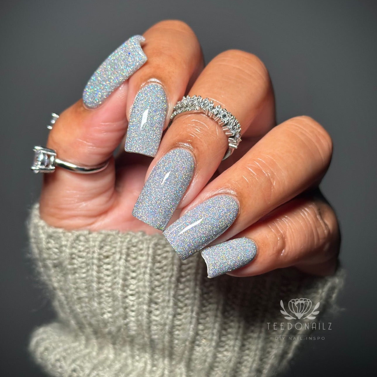 Hand with glittery silver nails wearing two rings, against a dark background