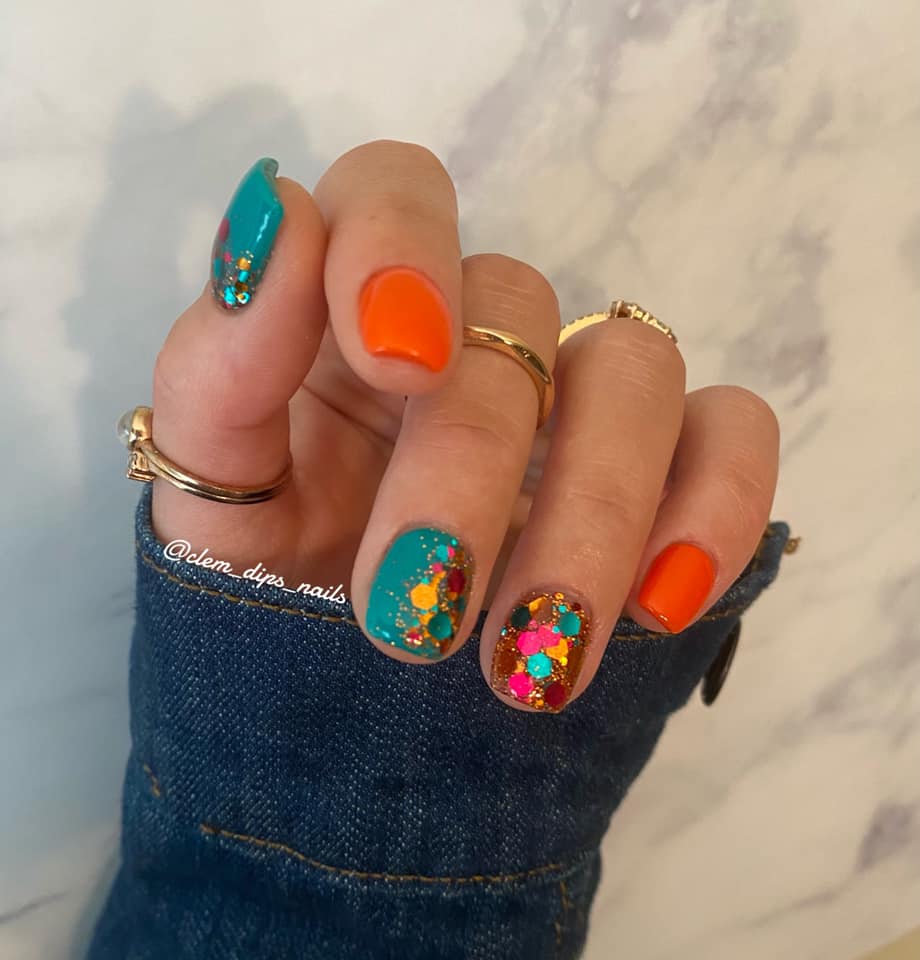 Teal and bright orange nails with chunky multicolor glitter accents on denim-sleeved hand with gold rings.