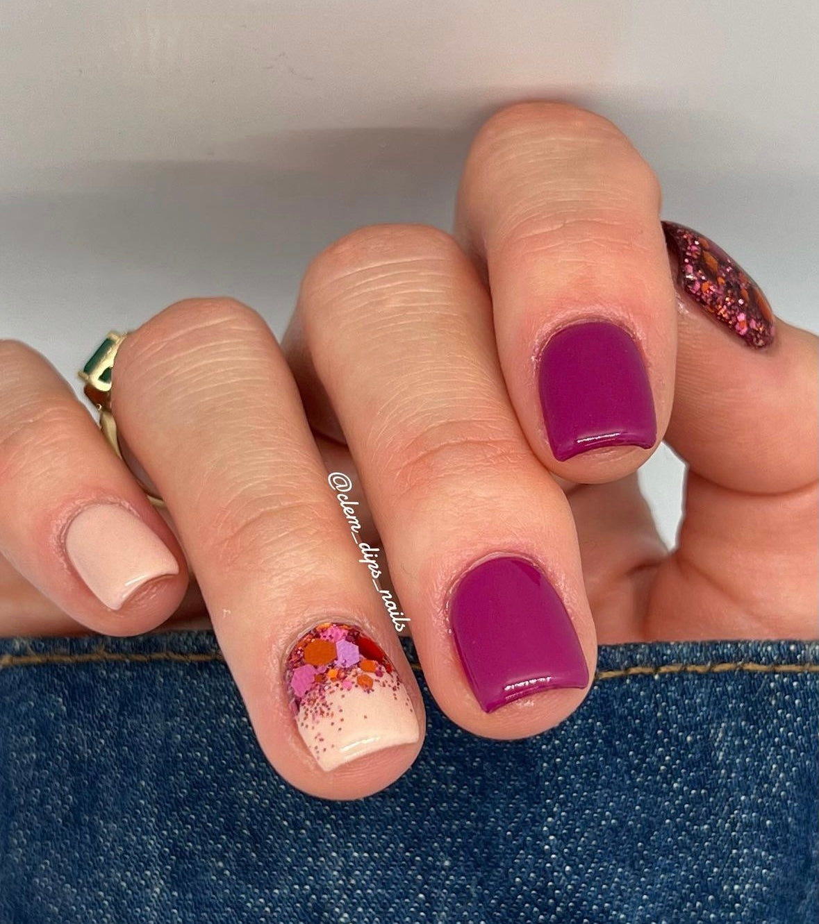 Hand with nails painted in solid nude, deep berry, and nude with orange and pink glitter accents on short square nails.