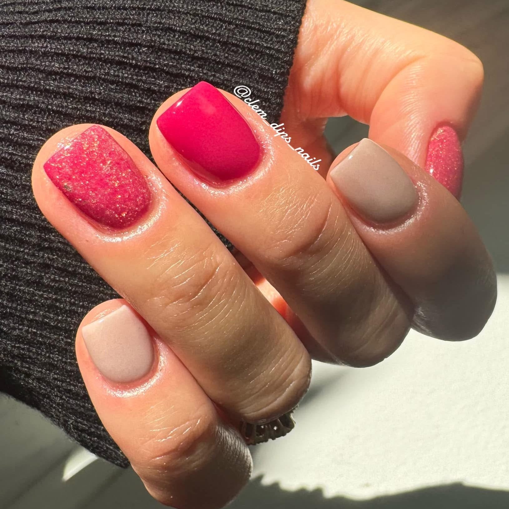 Hand with nails painted in pink glitter, glossy bright pink, and matte nude tones against dark ribbed fabric background.