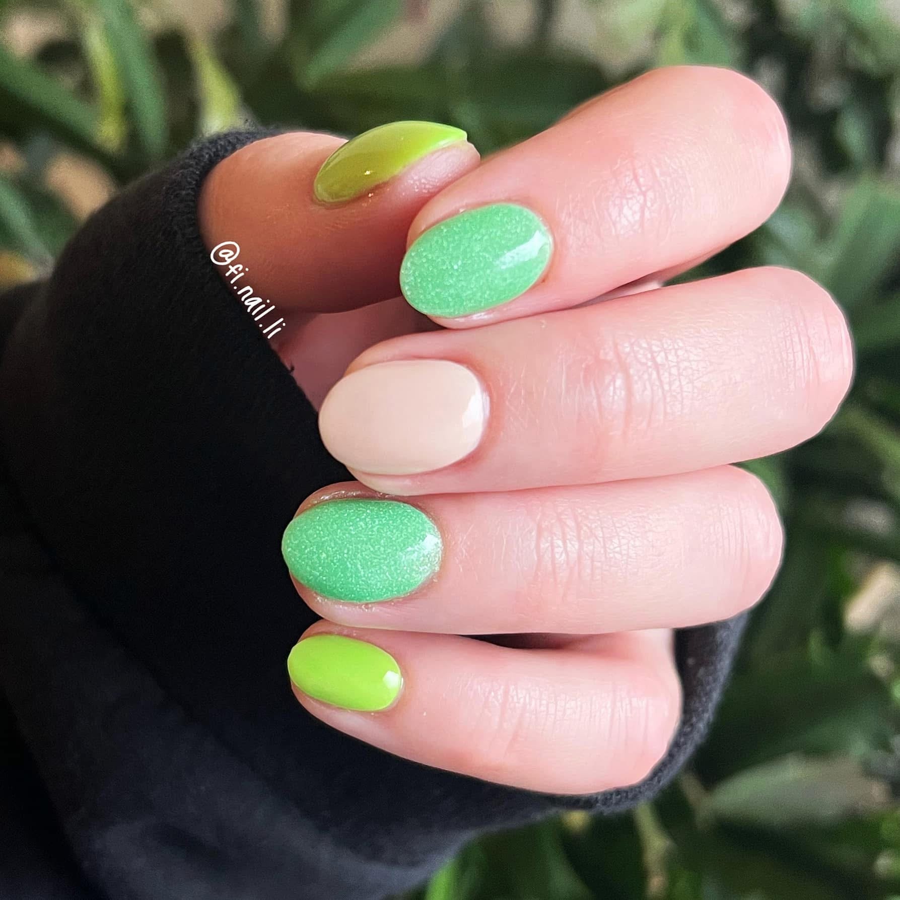 Hand with nails painted in mint green glitter, solid lime green, and soft beige polish against a blurred plant background.