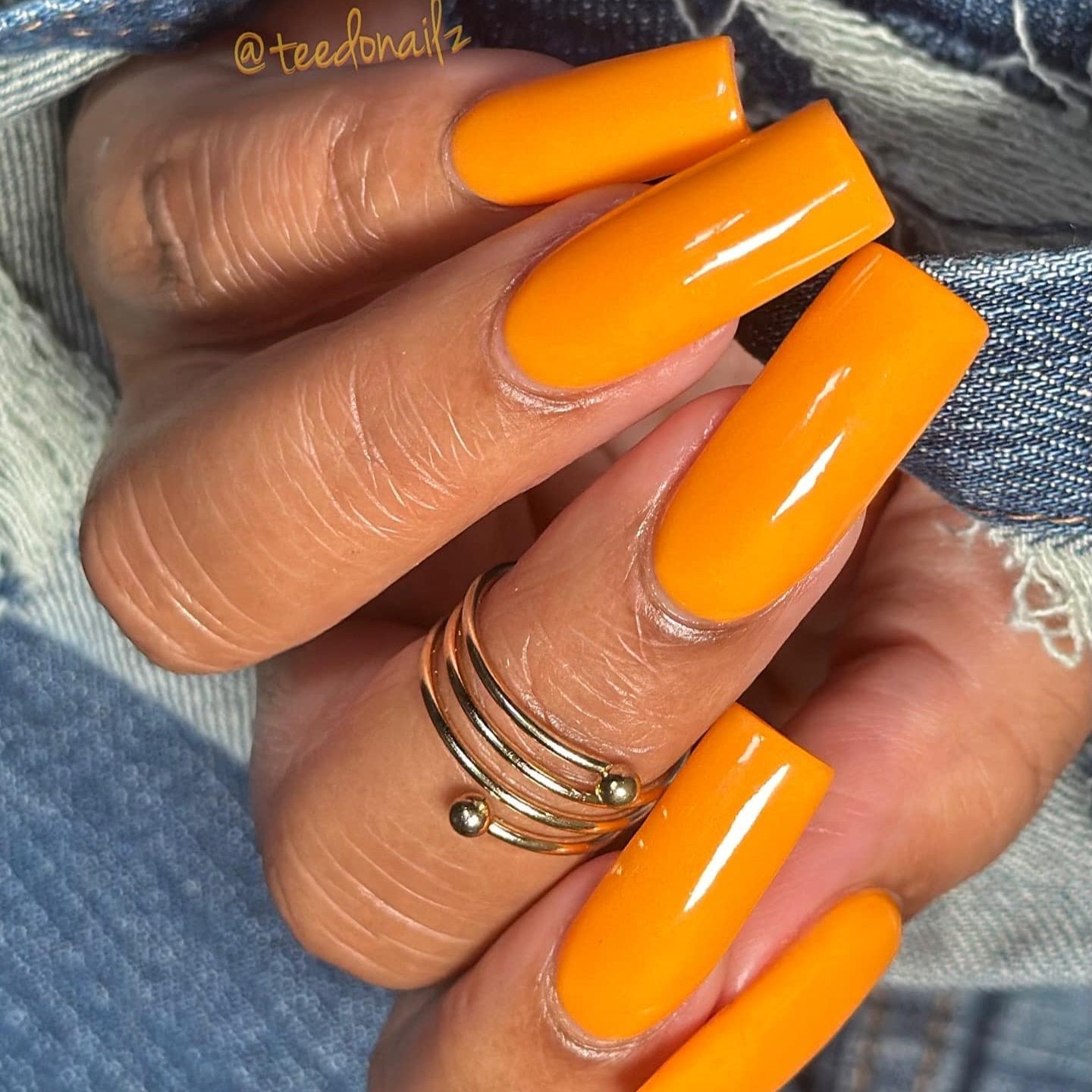Bright orange glossy long coffin-shaped nails with a smooth finish and a gold spiral ring on the finger.