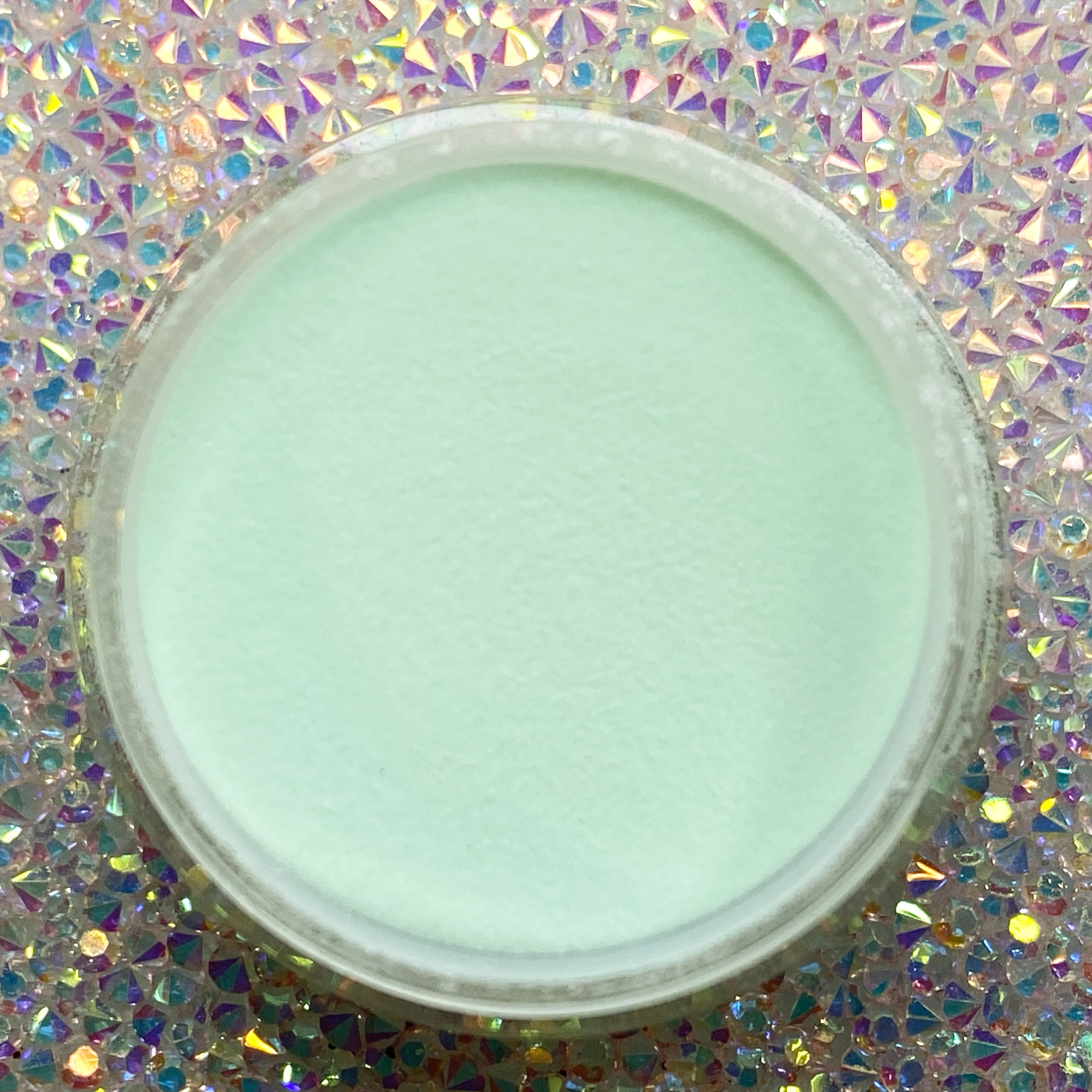 Small open container of smooth, light blue solid dip powder on a holographic surface.