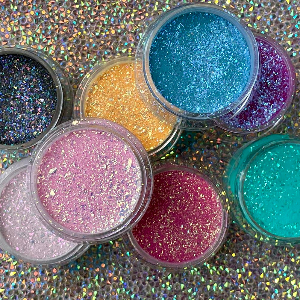 Assorted small jars of colorful glitter powders in pink, blue, green, gold, and purple shades on holographic background.