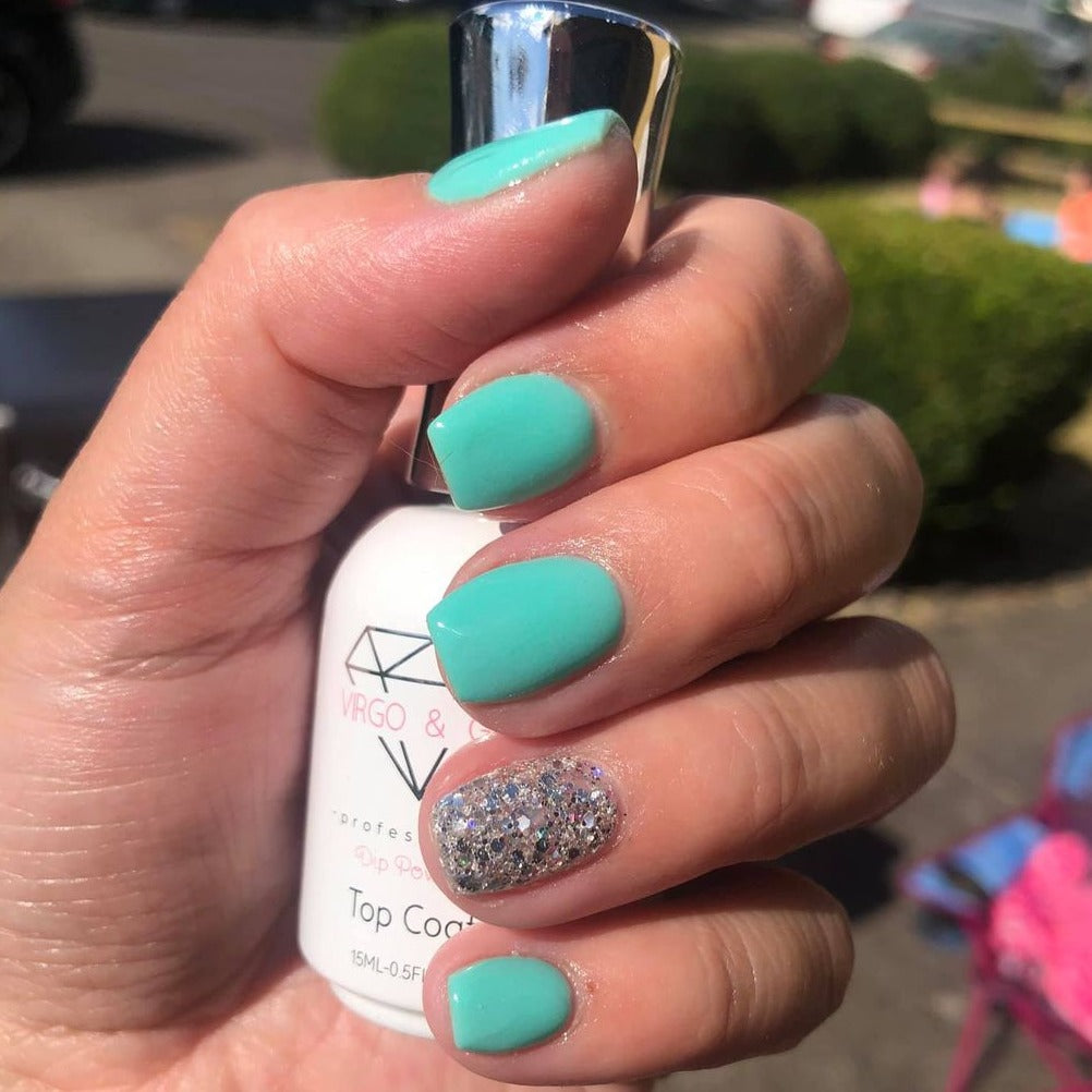 Hand with teal-blue matte nails and one accent nail coated in chunky silver glitter holding a white nail polish bottle.