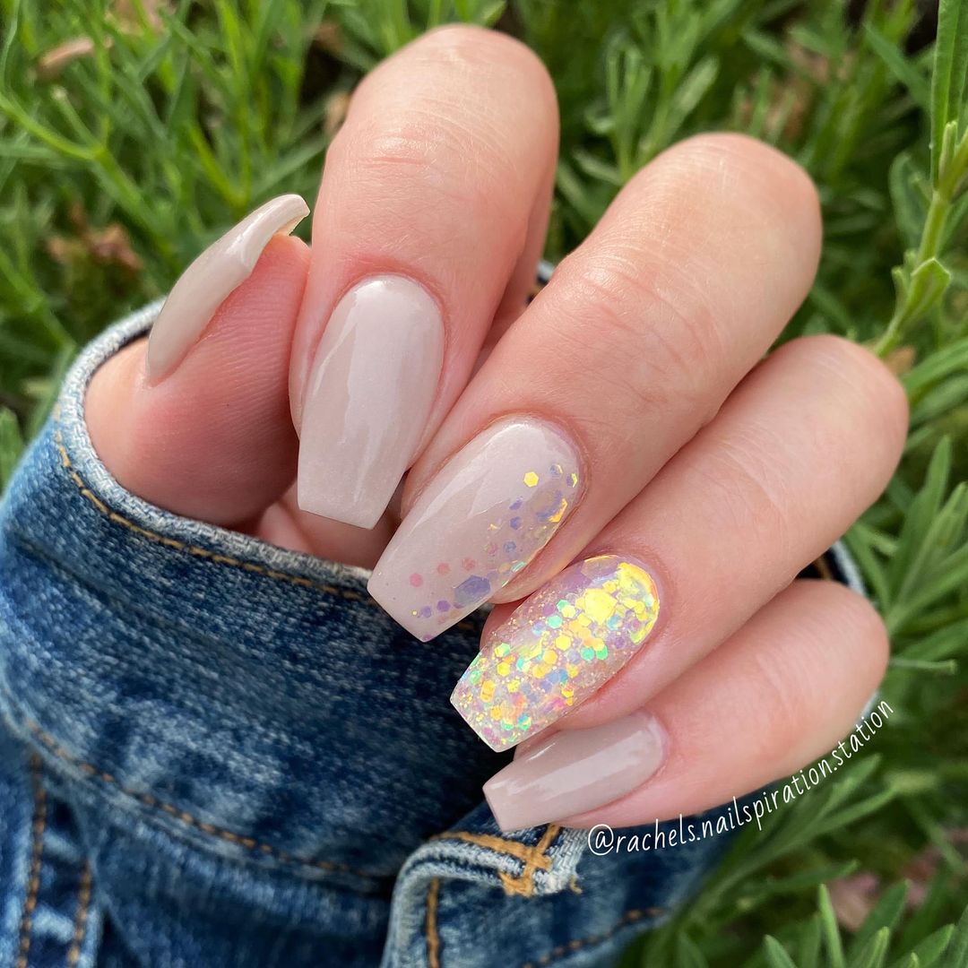 Hand with beige nude dip powder nails, one nail featuring iridescent glitter flakes, outdoors background.