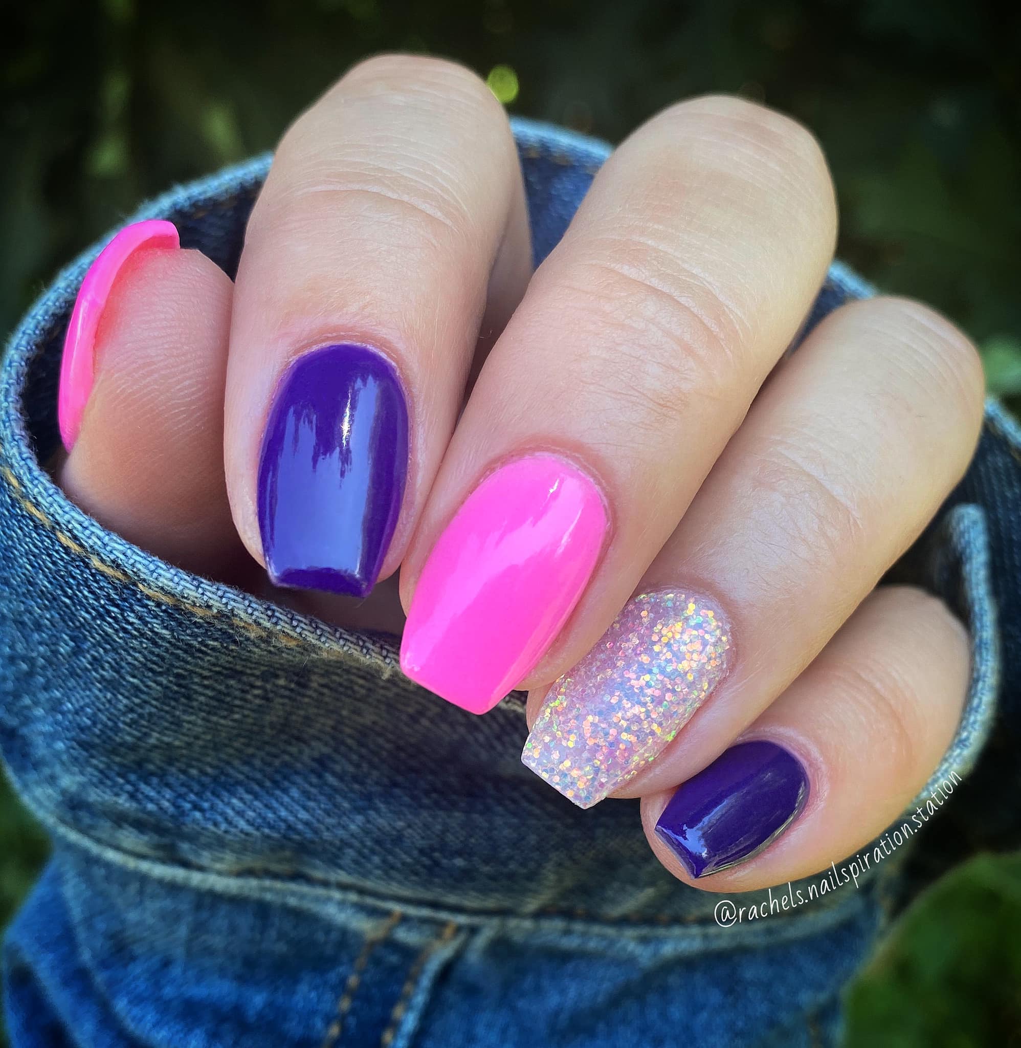 Fingernails with purple, bright pink, and glittery silver dip powder polish on a hand partially covered by denim fabric.