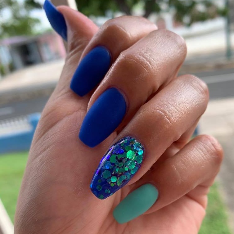 Matte deep blue nails with one teal matte nail and one accent nail featuring chunky blue and green glitter on glossy base.