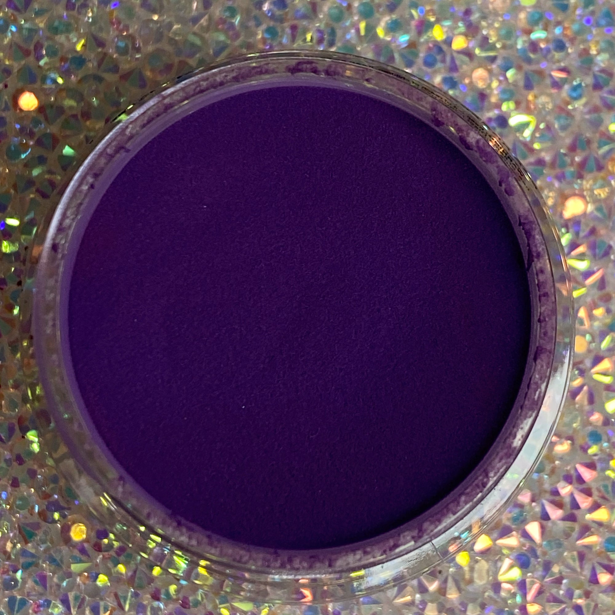 Open jar of deep purple solid dip powder with holographic surface beneath it.