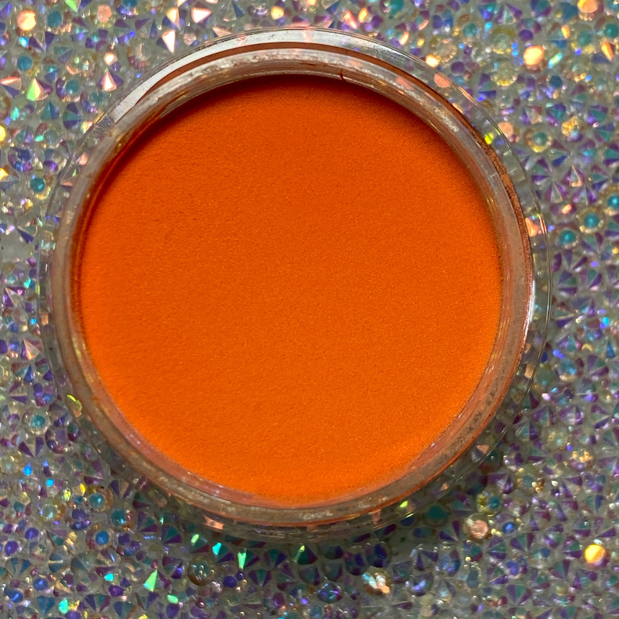 Bright solid orange dip powder in clear round jar on sparkling holographic surface.