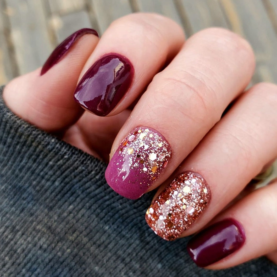 Hand with glossy deep burgundy nails, accent nails featuring chunky rose gold and pink glitter on pink base.