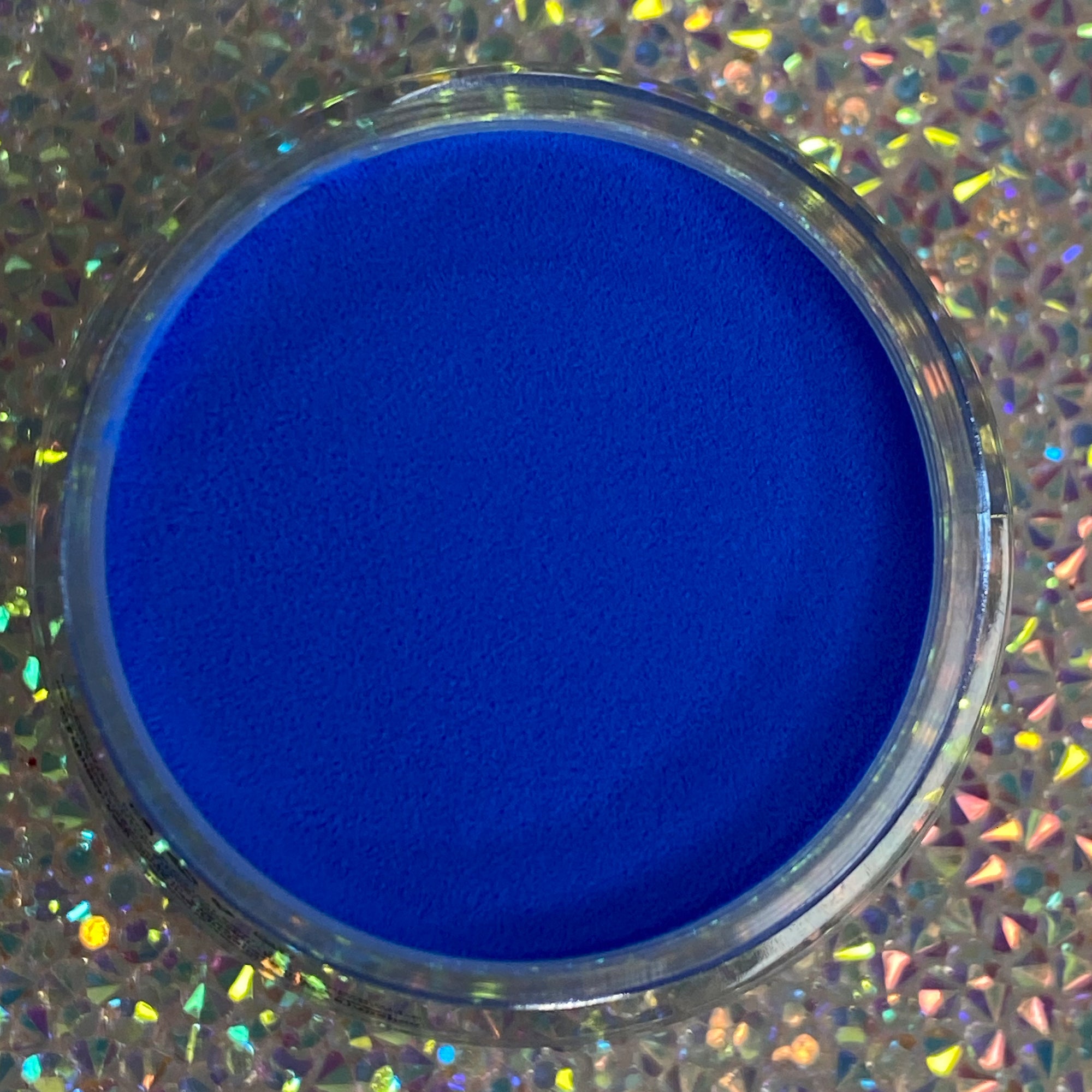 Bright cobalt blue dip powder in clear round container on holographic surface.