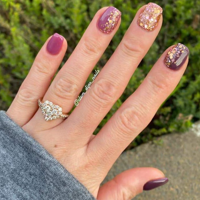 Hand with nails painted in deep purple and wine tones, gold glitter accent nails, adorned with a gold and diamond ring.