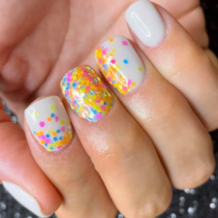 Short nails with pale pink polish, multicolored glitter accent nails featuring yellow, pink, blue, and purple dots.