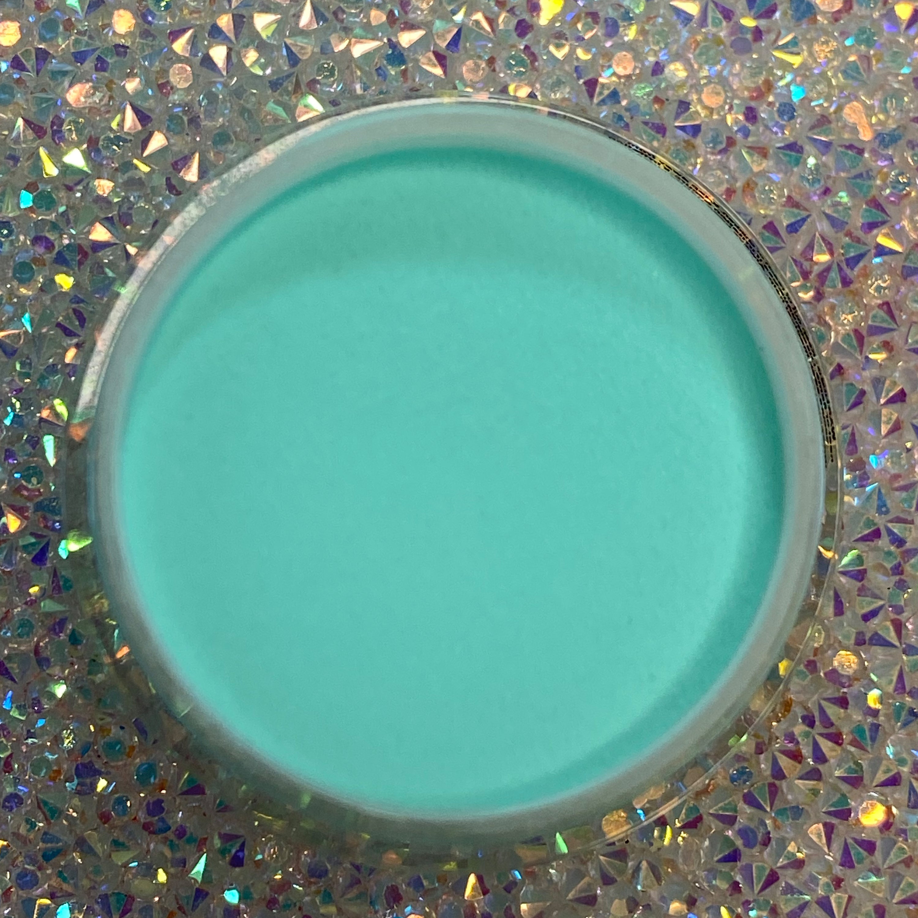 Round container of smooth teal dip powder nail polish on iridescent holographic background.