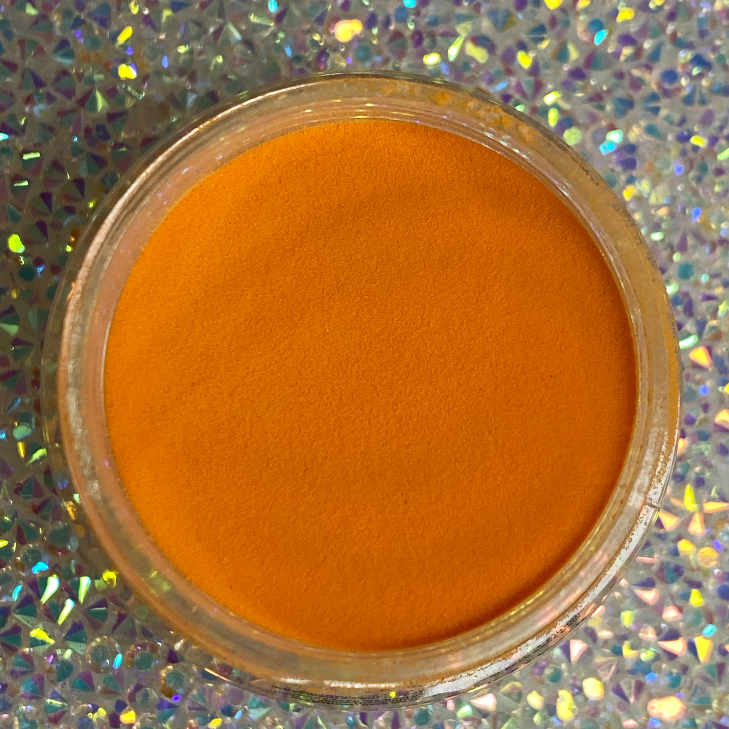 Bright orange dip powder in clear round container on holographic background.
