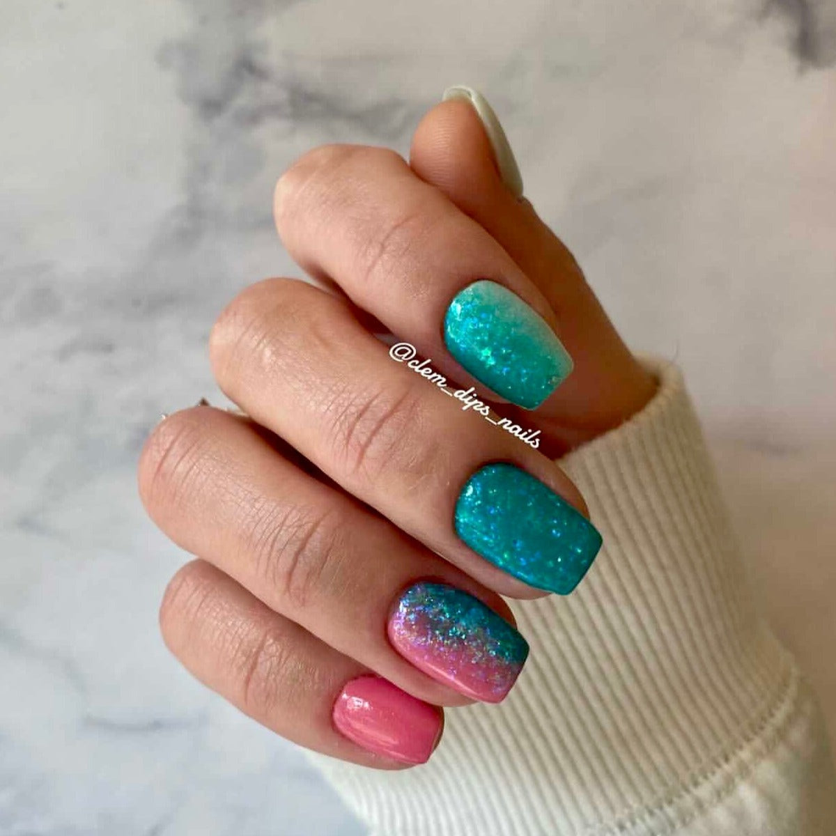 Hand with teal and pink glittery chameleon flake powder nails, short square shape.