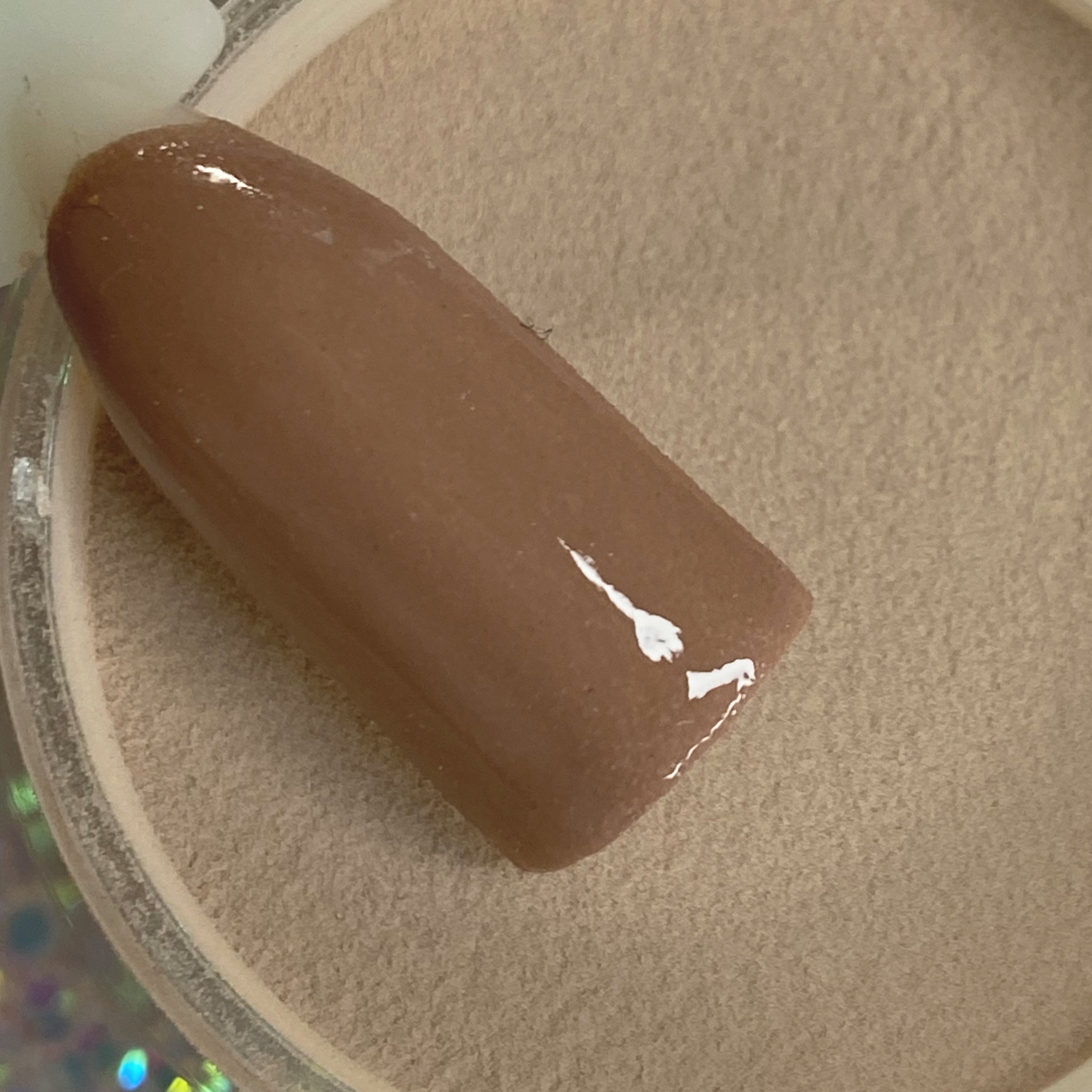 Smooth glossy medium brown nail tip resting on tan dip powder surface.
