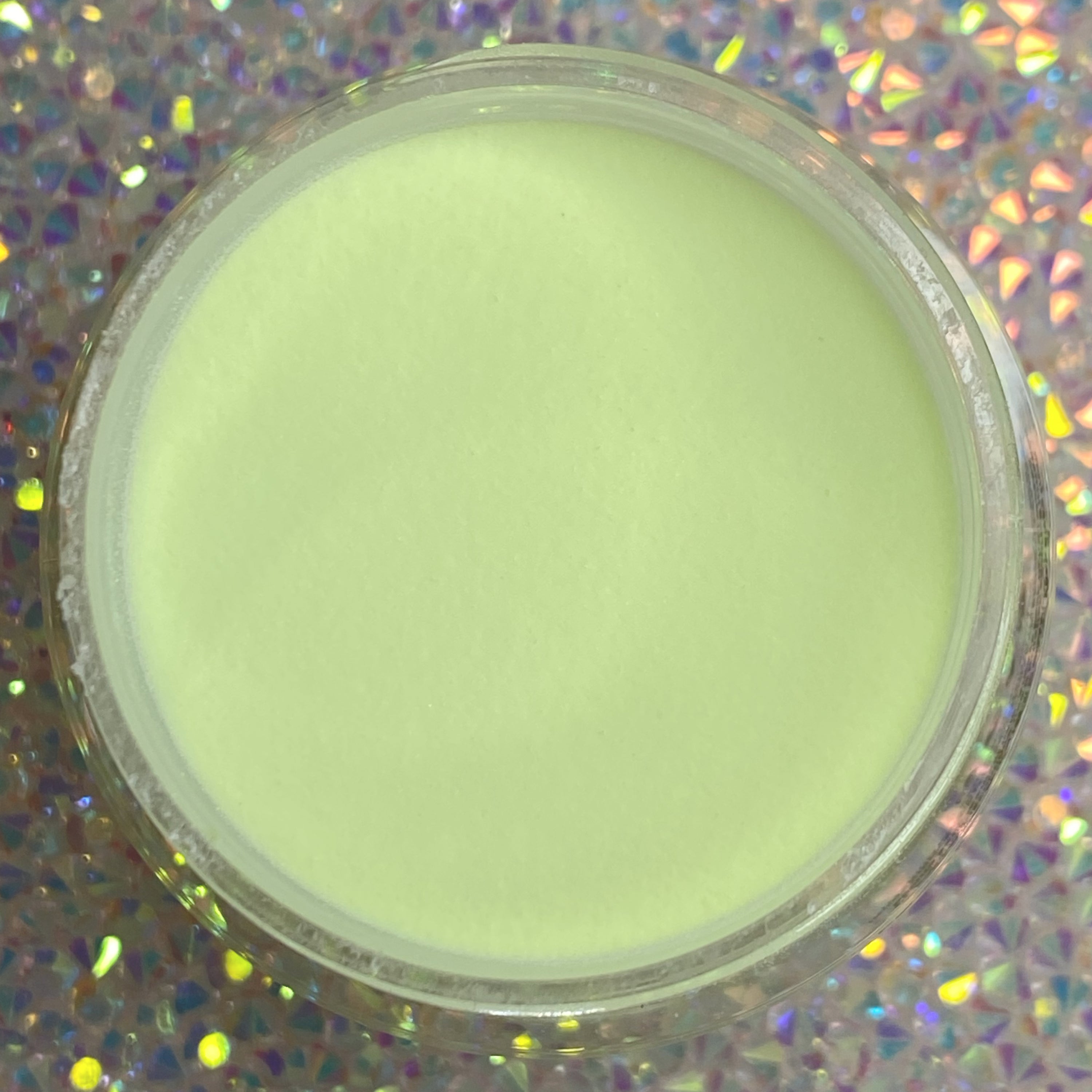 Clear container of smooth, pale pistachio green dip powder on holographic surface.