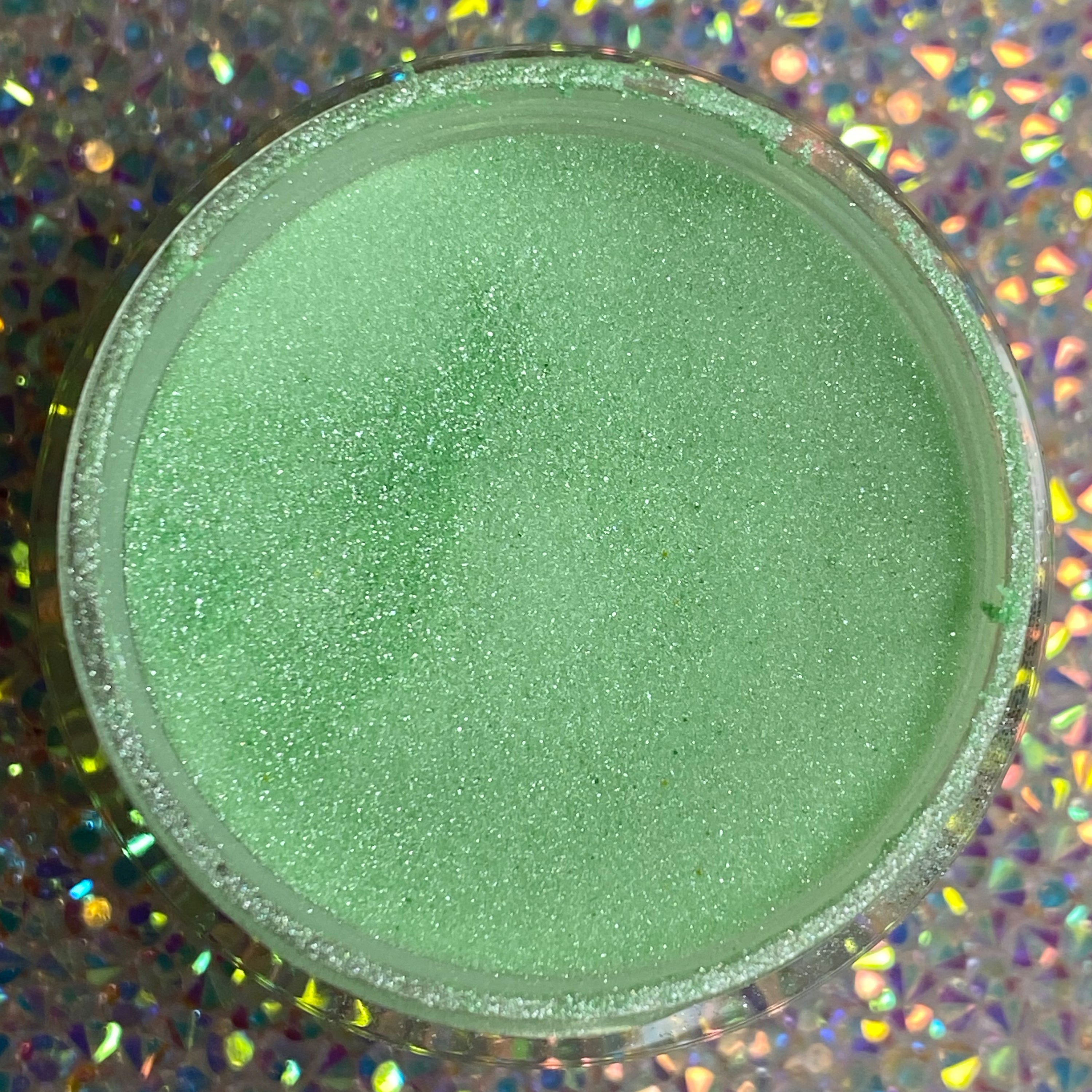 Open jar of fine mint green glitter dip powder with subtle shimmer on holographic background.