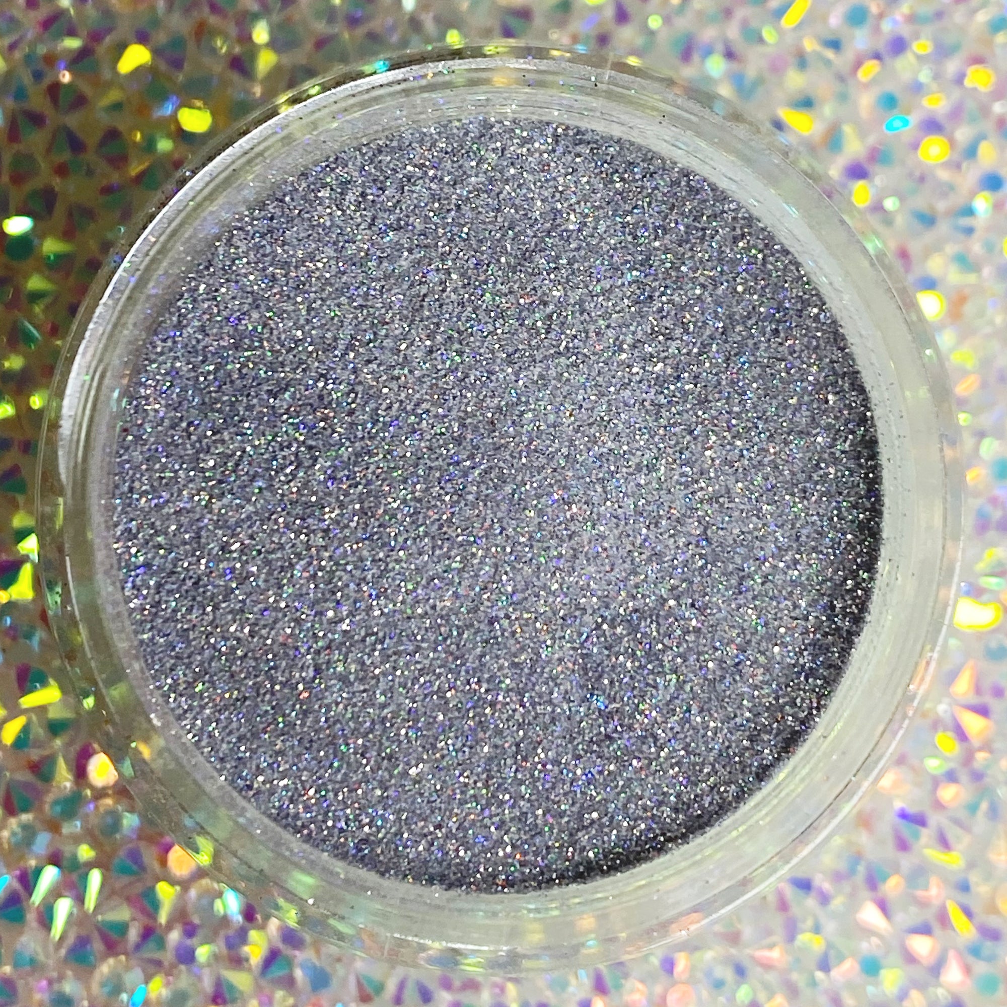 Fine holographic silver-gray glitter dip powder in a round clear container on holographic surface.