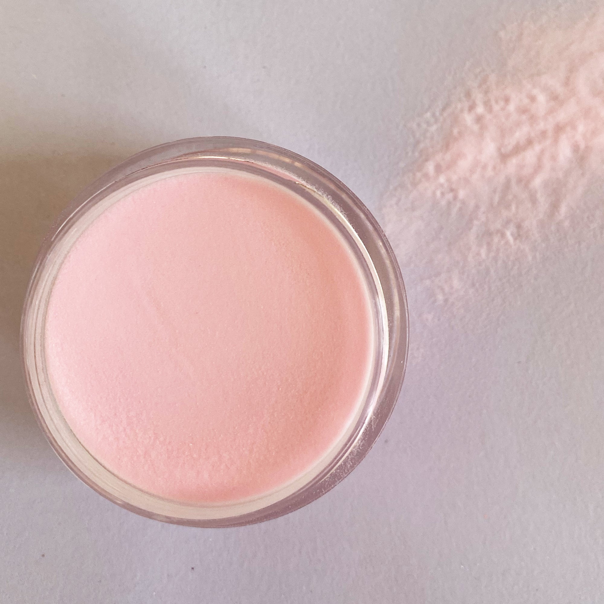 Round container of soft pastel pink dip powder with loose powder spilled nearby on light surface.