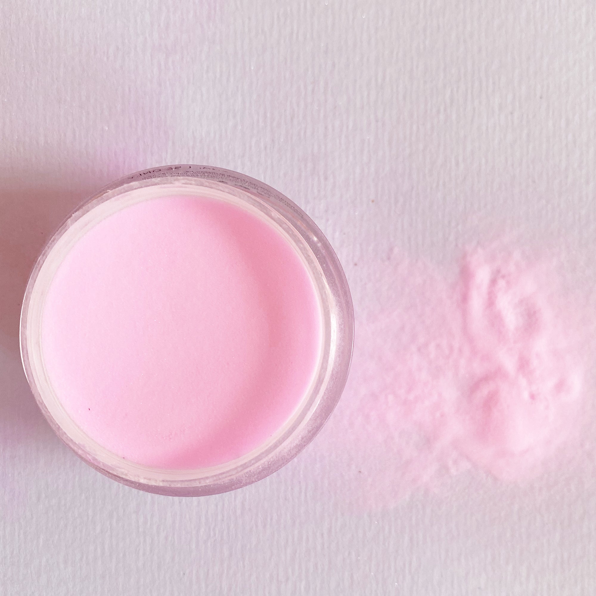 Open jar of soft pastel pink dip powder with loose powder spilled beside it on white surface.