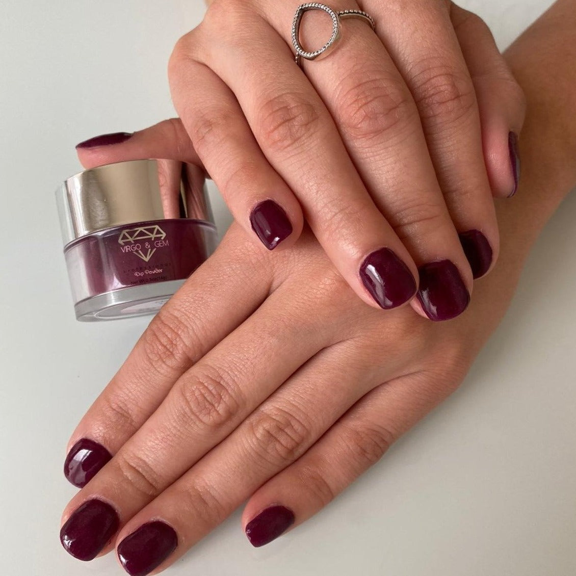 Hands with glossy deep wine purple nails next to a small jar of matching dip powder polish.