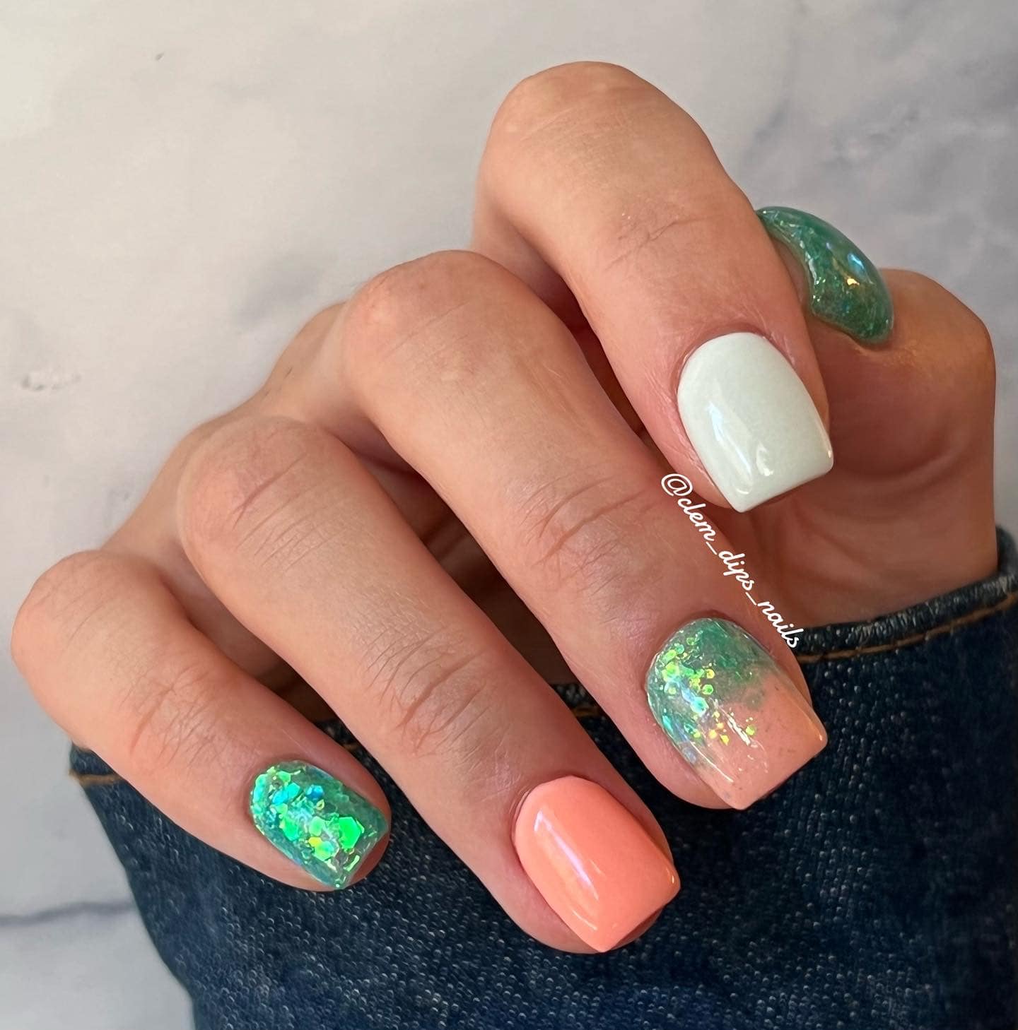 Hand with five acrylic nails in mint green glitter, solid white, coral orange, and iridescent shimmer designs.