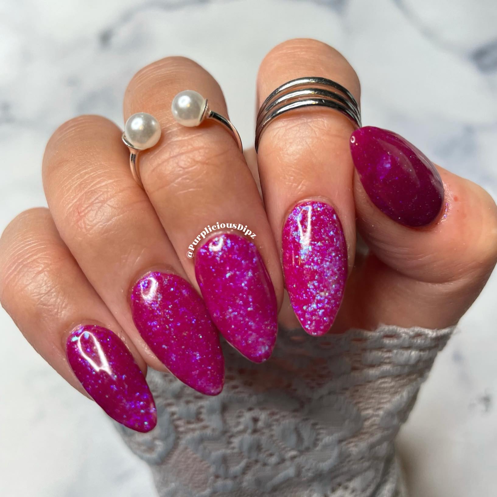 Bright purple almond-shaped nails with multicolor chamleon flake glitter on a hand with silver rings and pearl accents.