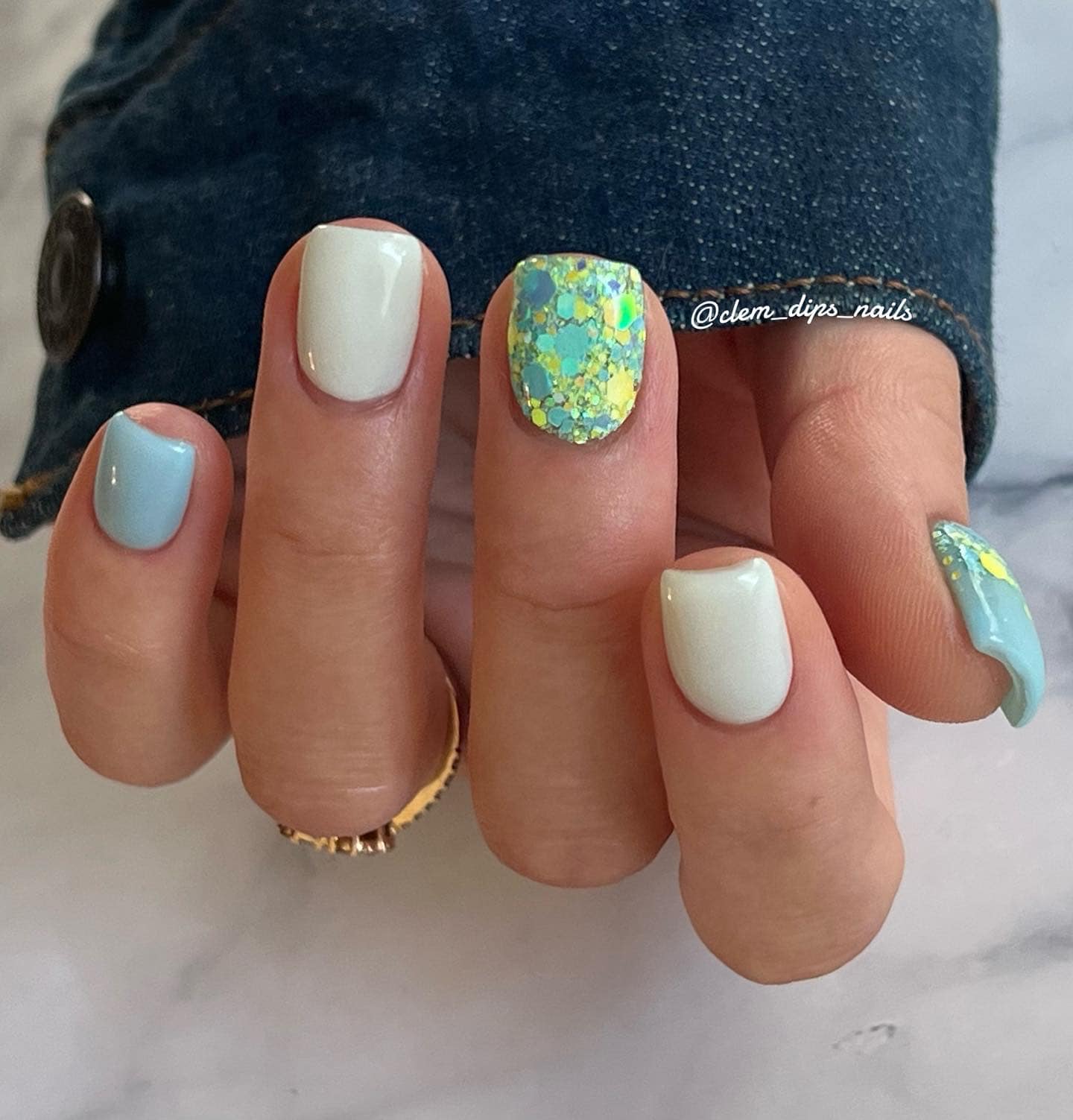 Short square nails with pastel blue and white polish, one nail with chunky green and blue glitter overlay.