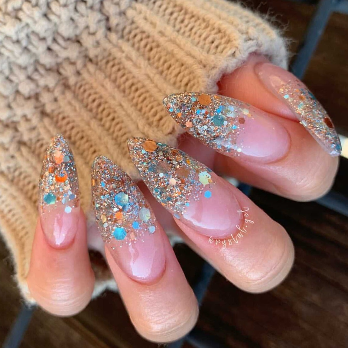 Long almond-shaped nails with clear base and chunky multicolor glitter in blue, copper, and silver tones.
