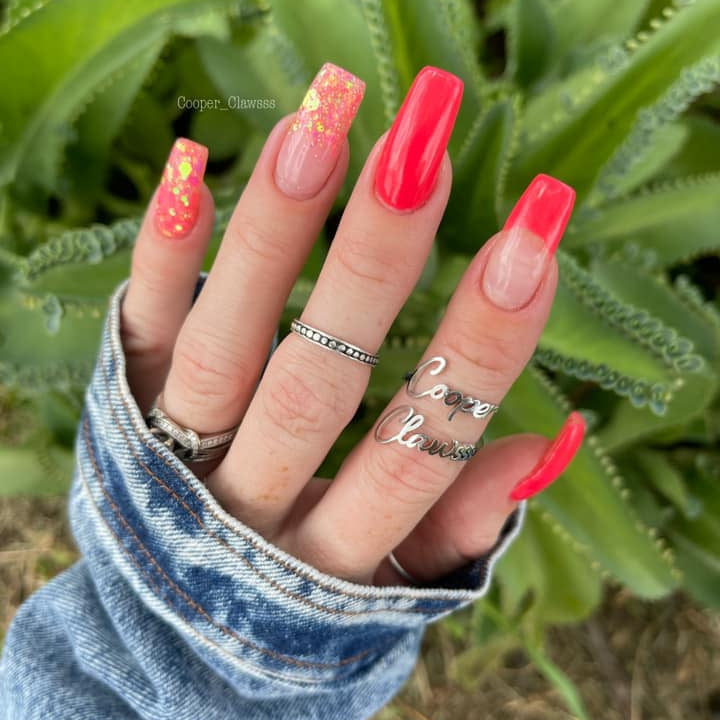Bright coral pink coffin nails with gold foil accents on natural nails with silver rings and denim sleeve.