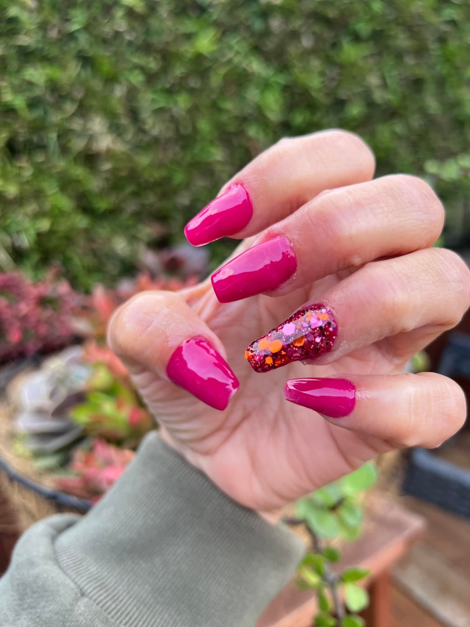 Hand with hot pink solid dip powder nails and one accent nail with red and orange glitter design outdoors.