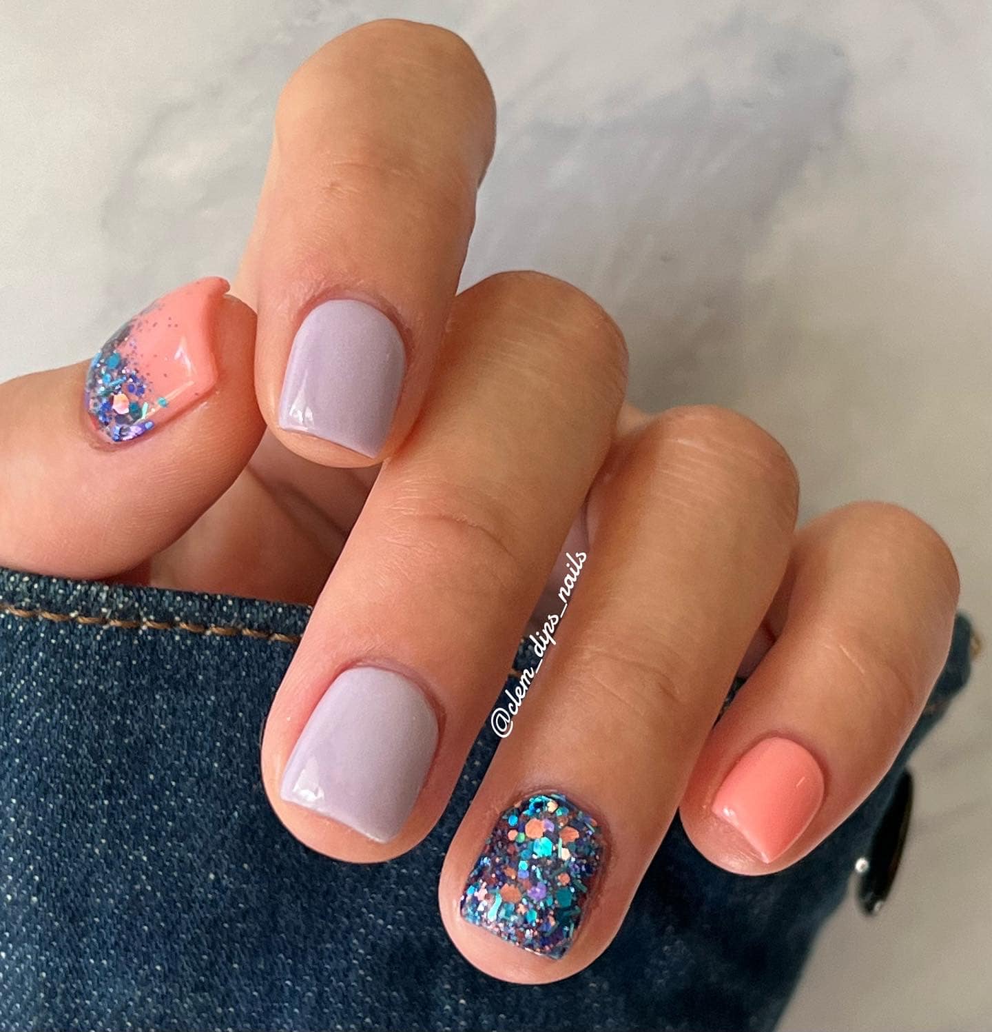 Short square nails with lilac purple polish on three fingers, coral pink on two, and one accent nail covered in blue and.