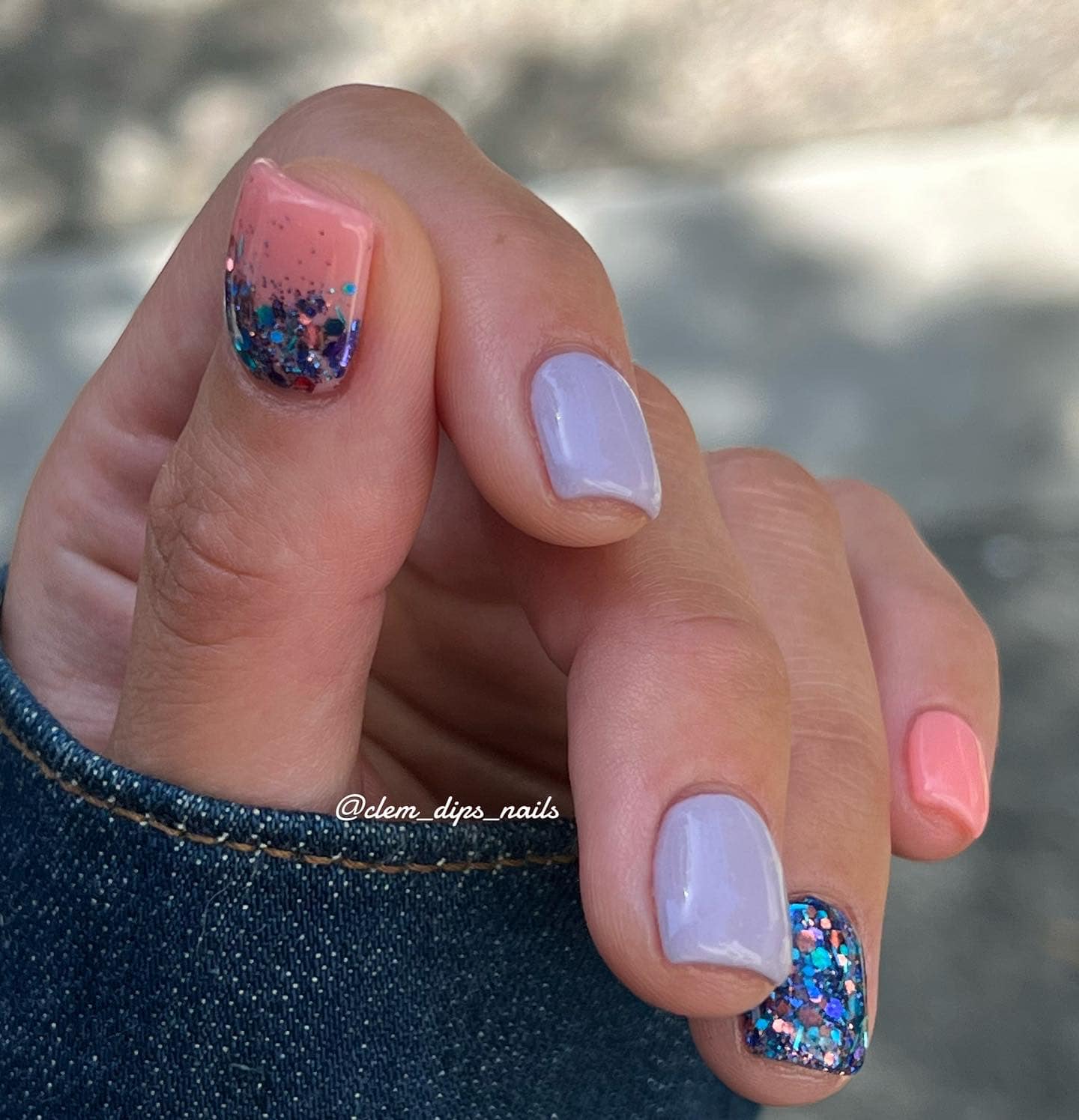 Hand with lilac and peach dip powder nails, two nails accented with chunky blue and purple glitter.