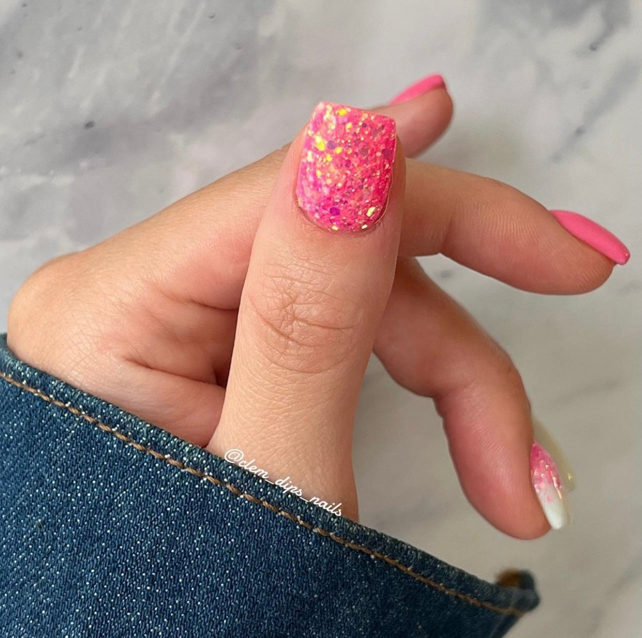 Pink chunky glitter nail polish with iridescent gold flakes on thumbnail and solid pink nails on other fingers against grey.